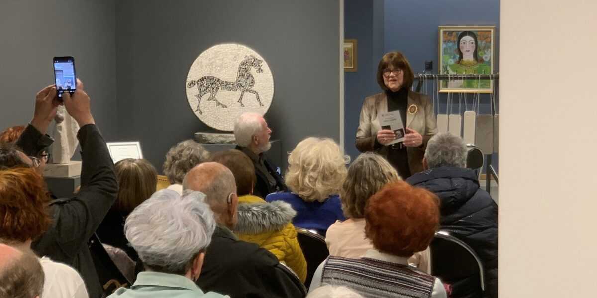 Odprtje razstave Odtisi v kamnu Odprtje razstave Odtisi v kamnu v Galeriji Trebnje. Veliko ljudi sedi na stolih, umetnica Jožica Škof stoji in nagovarja ljudi.