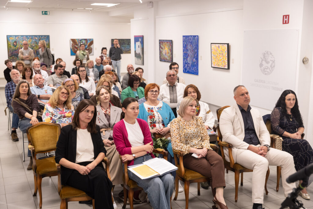 Ljudje sedijo na stolih v galeriji v prostorih Galerije Trebnje ob dogodku otvoritve razstave Marie Kozyrenko: Kjerkoli si. Foto: Boštjan Pucelj