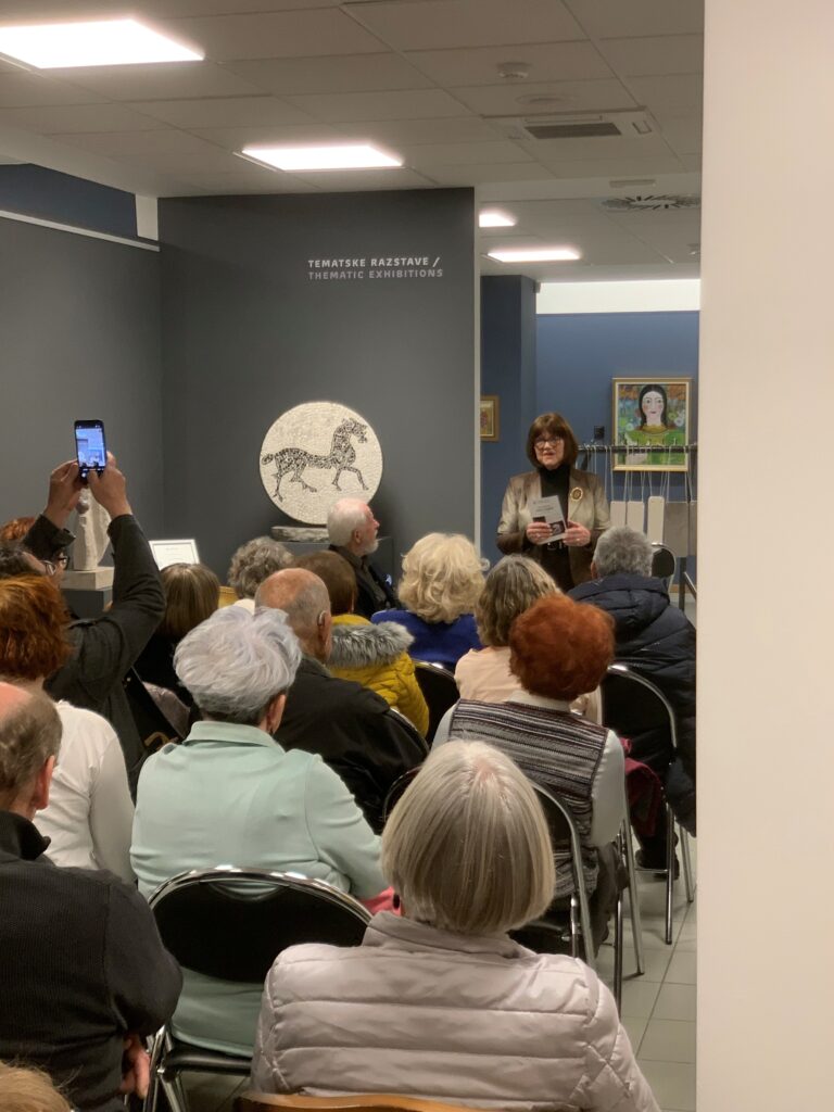 Odprtje razstave Odtisi v kamnu v Galeriji Trebnje. Veliko ljudi sedi na stolih, umetnica Jožica Škof stoji in nagovarja ljudi.