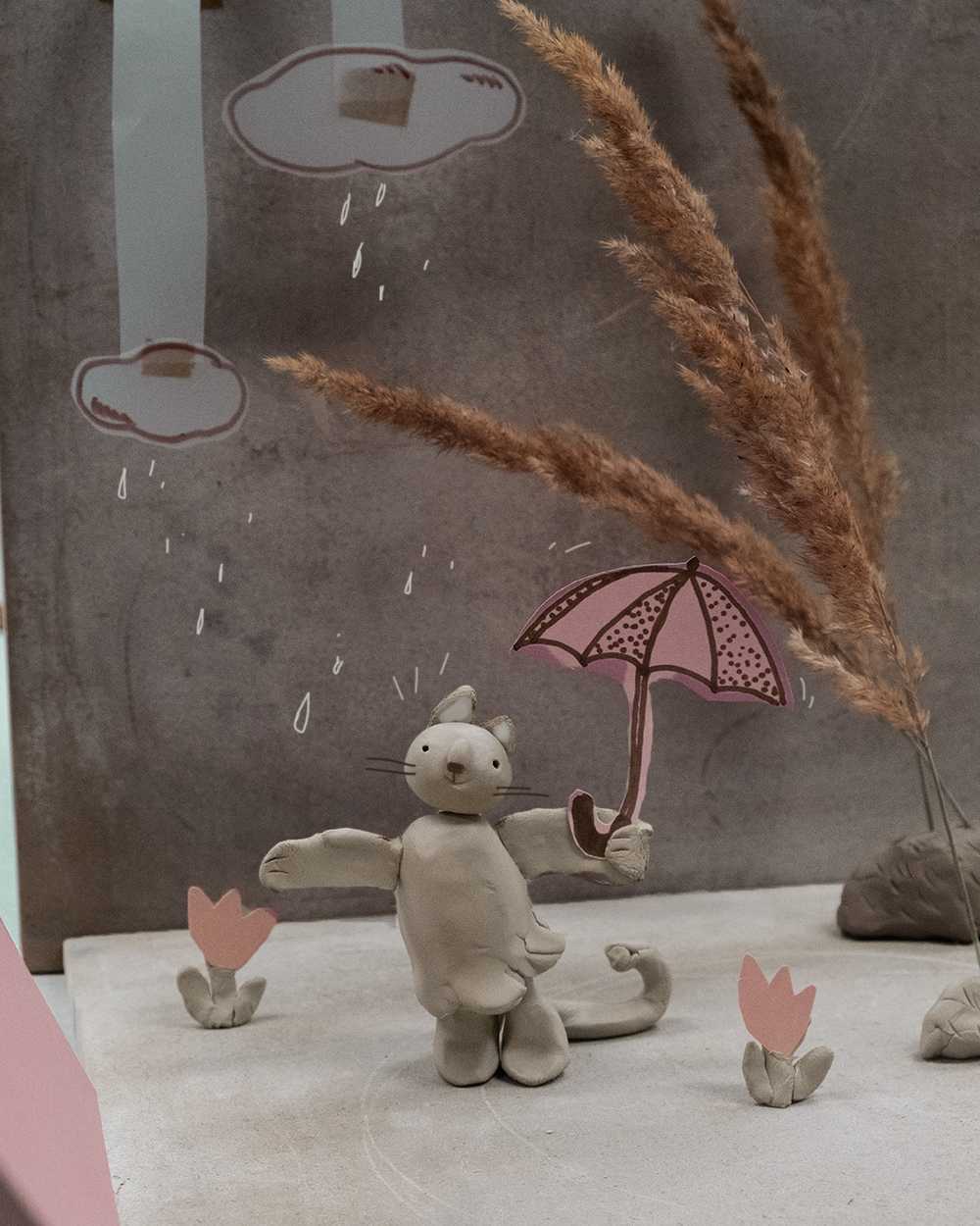 A clay cat. Photo: Shrusko A cat made of clay is holding a paper umbrella; above it are paper clouds and raindrops.